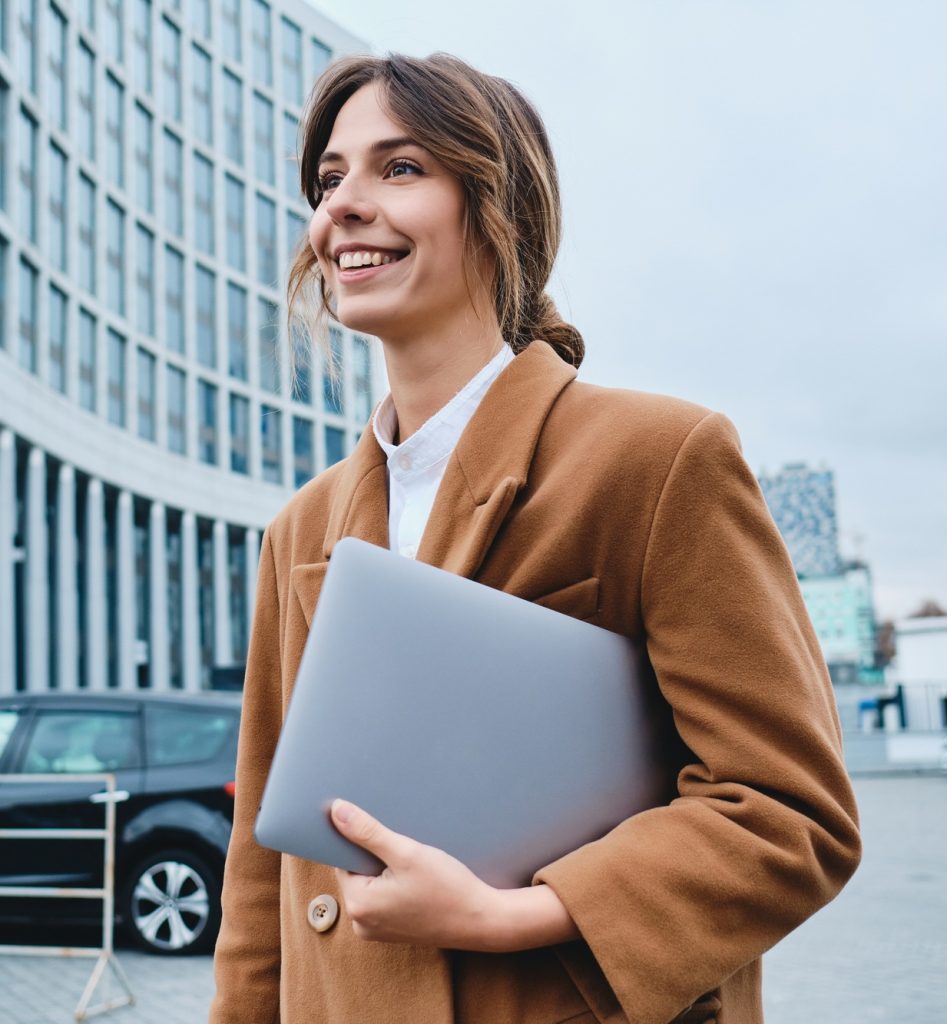 Young smiling lawyer in coat with laptop happily looking away walking through city street